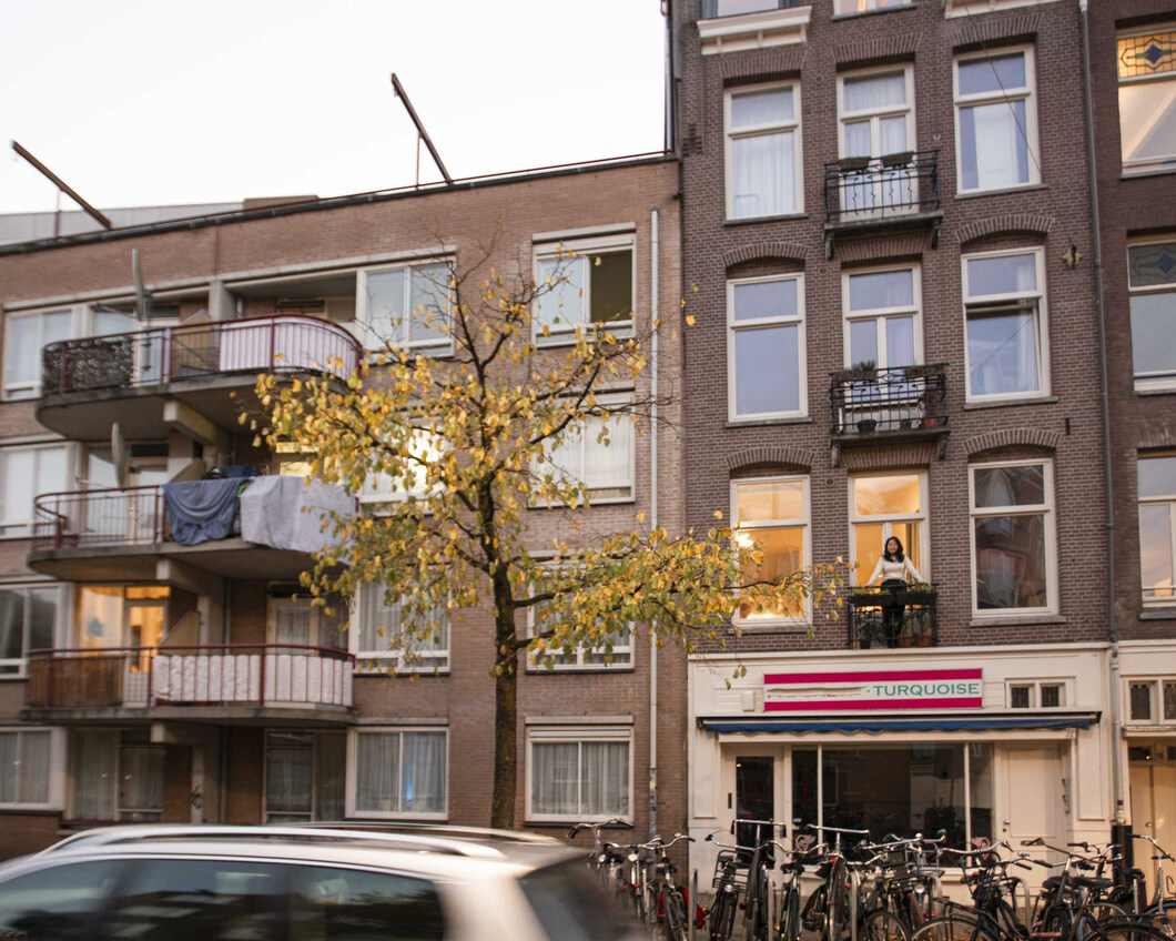 Het aangezicht van een aantal appartementen boven een buurtwinkel. Er staat iemand op het balkon. 