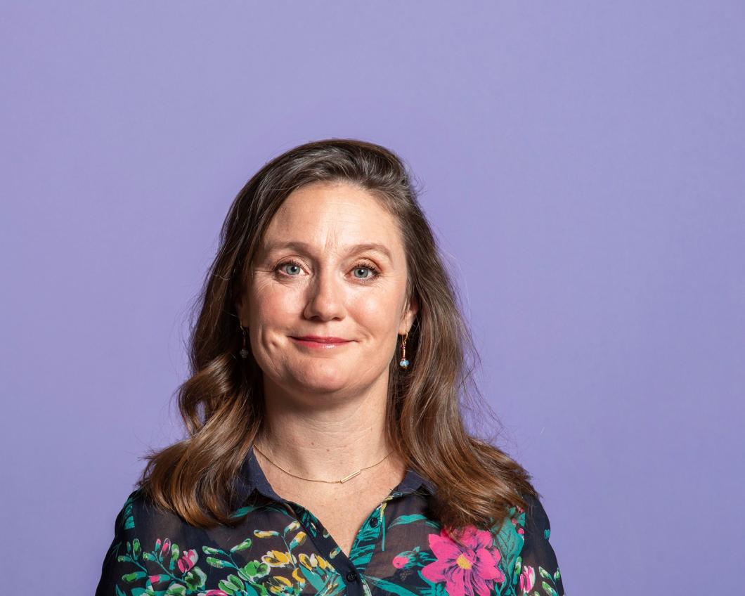 Foto van Suzanne Kröger, een witte vrouw. Ze draagt een zwarte blouse met groene en rode planten erop, heeft bruin haar en heeft een kleine glimlach.