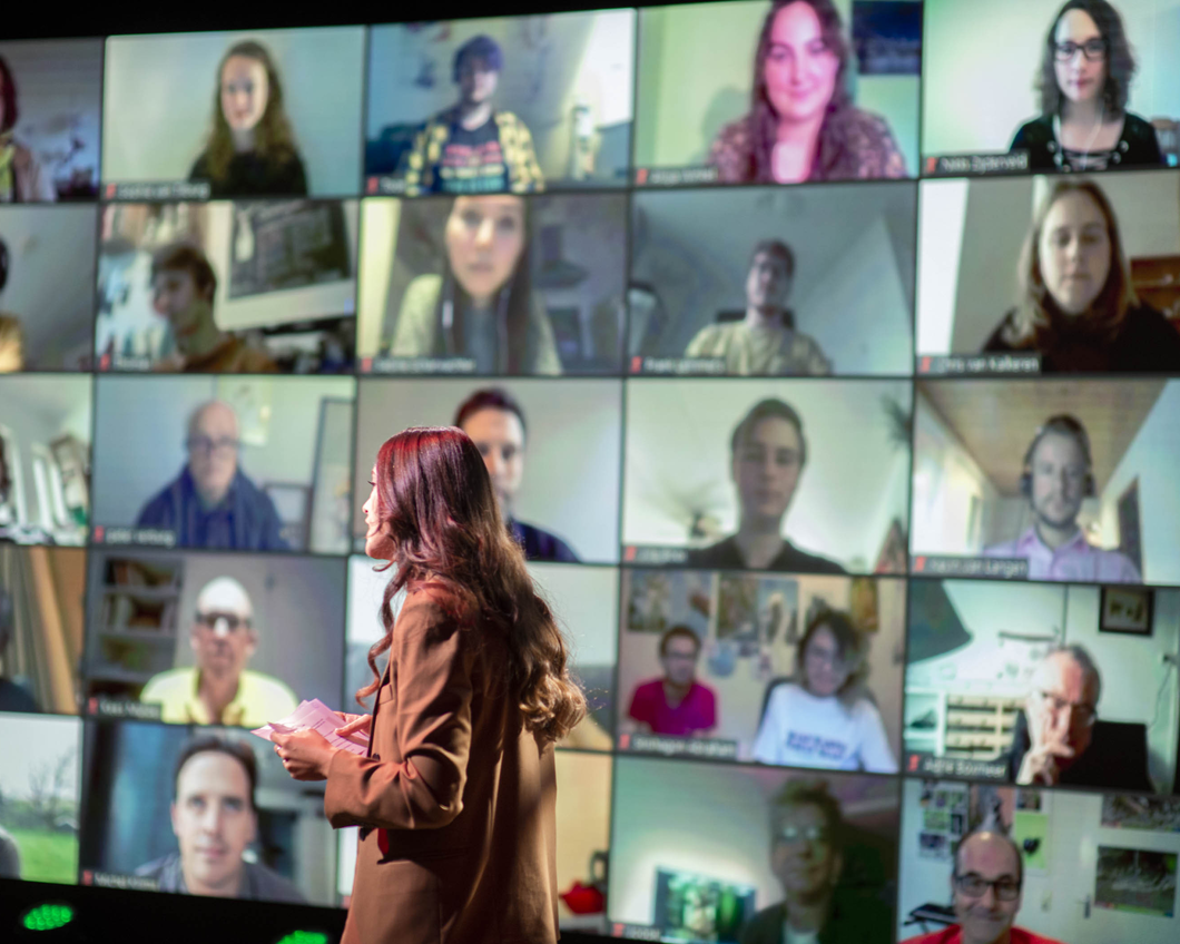 Emmelie, een vrouw met lang donker haar, staat voor een muur met een videoconferentie met allerlei mensen erop.