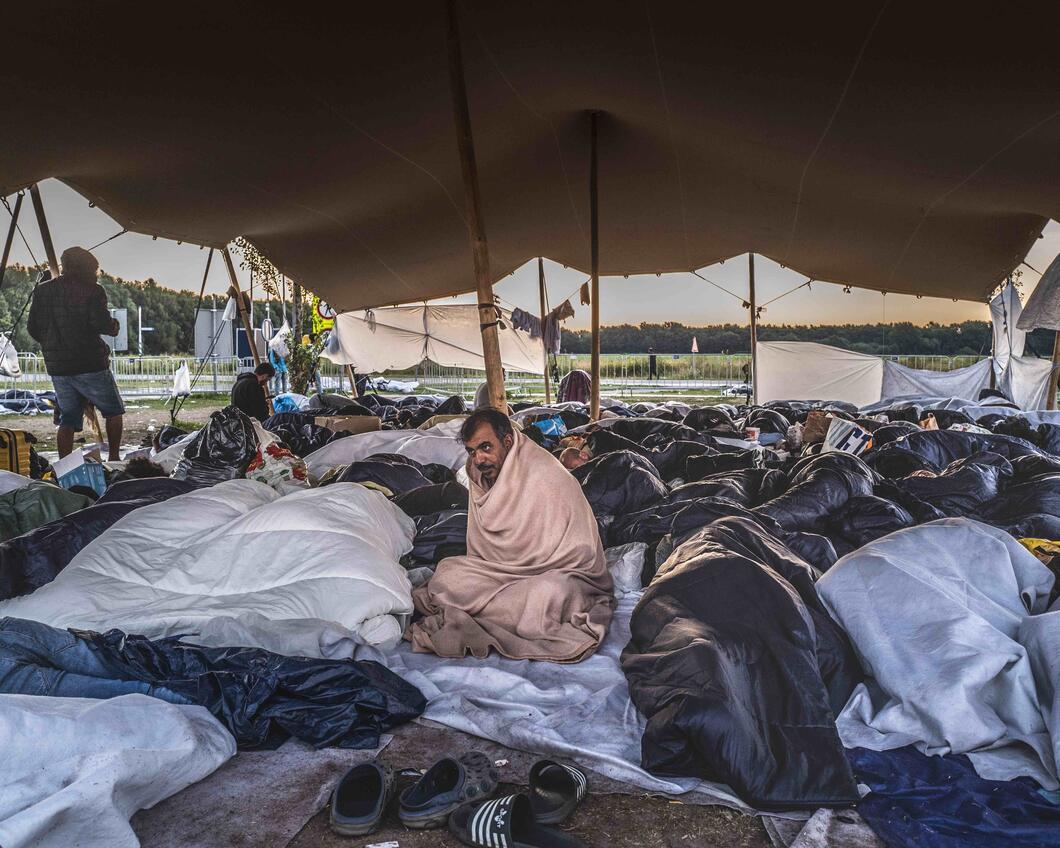 Een heleboel mensen liggen dicht op elkaar onder een zeil te slapen. Ze zijn vluchtelingen die liggen te wachten voor de poort van Ter Apel. In het midden zit een man, hij is gewikkeld in een deken en kijkt voor zich uit. 