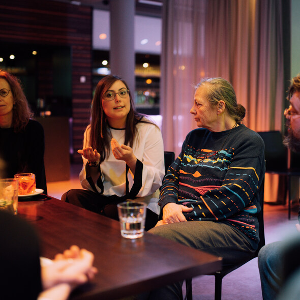Een jong persoon zit aan tafel, pratend met veel handgebaren. Een ouder persoon luistert. Om hen heen zitten nog meer luisterende mensen.
