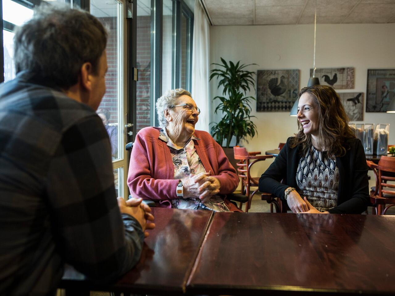 Een lachende oudere vrouw zit aan tafel met een jongere vrouw en een man